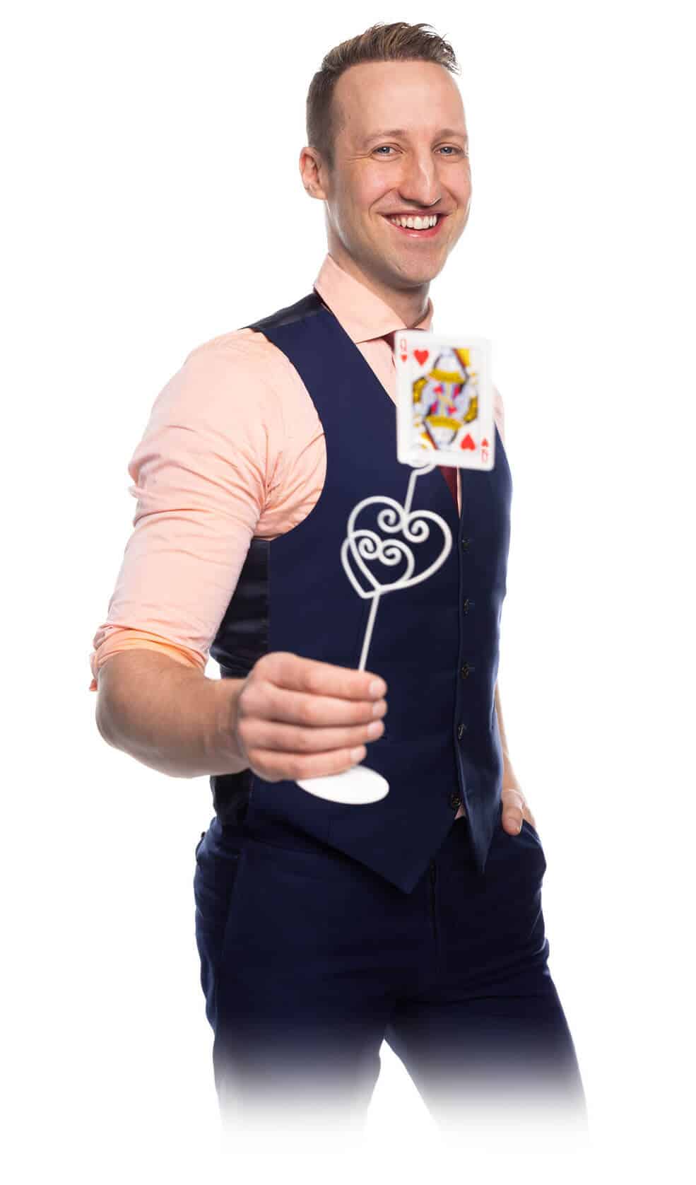 The wedding magician Fabian Schneekind smiles into the camera. In his hand, he holds a heart-shaped memo holder with a playing card clamped in it. The card is the Queen of Hearts and plays a crucial role in his wedding show.